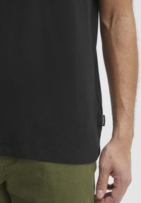 Camiseta de algodón negra con mangas cortas, cuello redondo y abertura lateral. Presenta una pequeña etiqueta en el dobladillo. Combinada con pantalones verde oliva.
