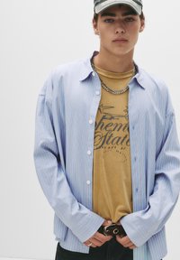 Stribet blå button-up skjorte over en tan grafisk t-shirt; har en kædehalskæde og mørke jeans med bælte. Glat tekstur, afslappet pasform.