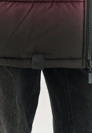 Prešívaná bunda s čiernym gradientným efektom. Má zapínanie na zips, šijacie detaily a malú textilnú nášivku na leme. Kombinovaná so sivými džínsami.