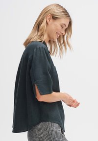 Blusa verde scuro a maniche corte con vestibilità ampia, spacchi laterali e taschino sul petto. Il tessuto testurizzato arricchisce il design complessivo.