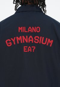 Tamsiai mėlynas švarkas su raudonais išsiuvinėtiais žodžiais: "MILANO GYMNASIUM EA7" ant nugaros. Lygus, lengvas audinys ir struktūrizuota apykaklė.