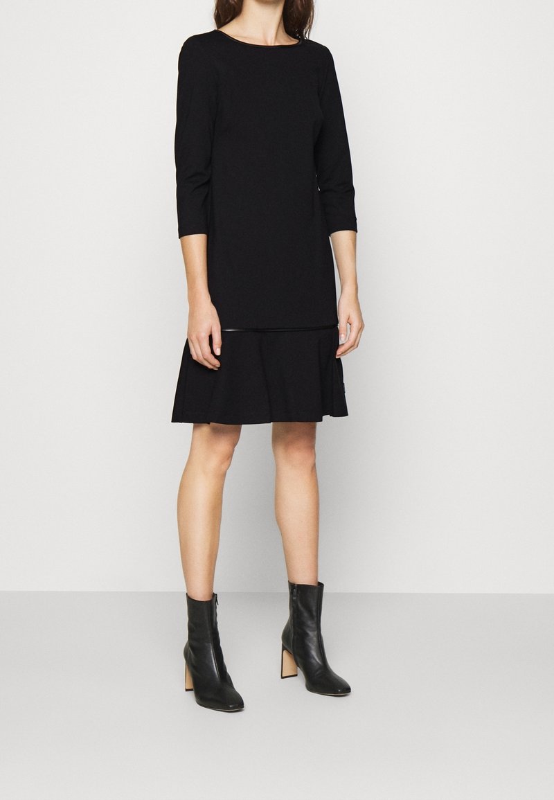 Femme portant une robe noire jusqu'au genou avec des manches trois-quarts et des bottines noires à talons blocs, debout devant un fond uni.