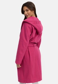 Accappatoio in pile di un vivace colore rosa, con cappuccio, cintura e un delicato dettaglio del logo sul retro, dotato di tasche frontali e una consistenza morbida.