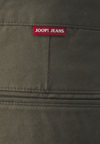 Tissu vert olive avec une texture lisse ; comporte une étiquette rouge cousue portant la mention "JOOP! JEANS." Design simple avec des lignes épurées et sans motifs visibles.
