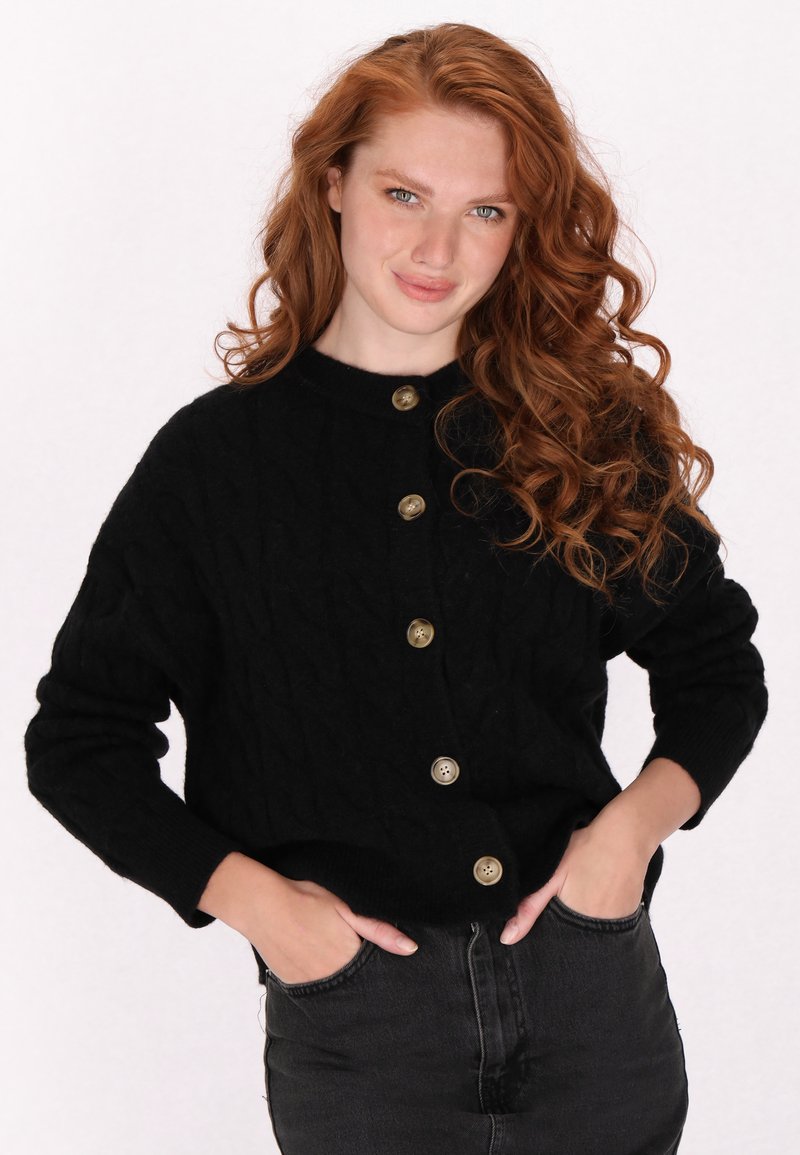 Zwarte gebreide cardigan met een getextureerd kabelpatroon, voorzien van grote gouden knopen en een ronde halslijn, gecombineerd met donkere jeans.