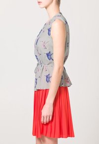 Bezramenný peplum top v světle šedé barvě se vzorem kolibříků, spojený s plisovanou červenou sukní. Hladká tkanina, přiléhavý silueta, boční pohled.