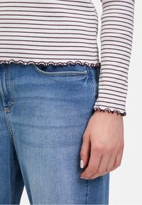 Gestreiftes Langarmoberteil mit einem gewellten Saum in Weiß und Bordeaux, kombiniert mit hellblauen Jeans. Hand sichtbar mit einem neutralen Nagellack.