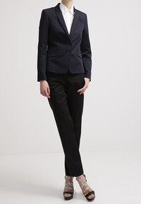Mujer vestida con un blazer negro a medida, camisa blanca con cuello, pantalones negros y sandalias de tacón alto con tiras de piel de serpiente, de pie sobre un fondo blanco.