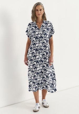 Vrouw met schouderlang grijs haar draagt een marineblauwe midi-jurk met witte bloemenprint en witte sneakers, staand tegen een effen muur.