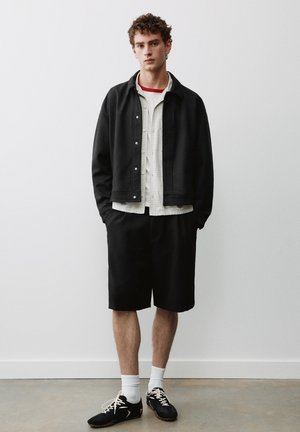 Ung mand der står op ad en hvid væg iført sort jakke, ternet skjorte, sorte shorts, hvide sokker og sorte sneakers med snørebånd.