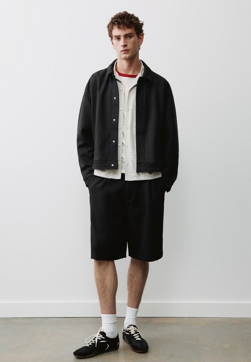 Jeune homme debout contre un mur blanc, portant une veste noire, une chemise à carreaux, un short noir, des chaussettes blanches et des baskets noires avec des lacets.