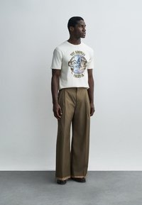 Álló férfi fehér, grafikával és szöveggel díszített pólóban, magasderekú, bő szárú barna nadrággal és sötét cipőben.