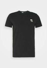 T-shirt nera in cotone con maniche corte, colletto rotondo e una piccola grafica a fumetto sul lato sinistro del petto, raffigurante un volto con occhiali.