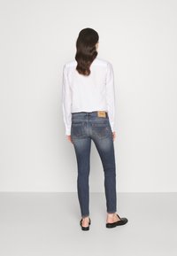 Camisa blanca de manga larga combinada con jeans ajustados azul oscuro. Los jeans tienen bolsillos traseros y una etiqueta marrón en la cinturilla. Calzado negro tipo slide.