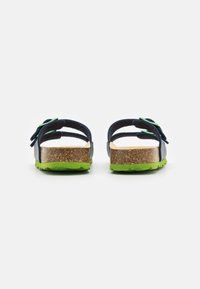 Två sandaler sedd bakifrån med en mörkblå ovandel, grön sula och korkfotbädd, med en dekorativ grön accent på remmen.