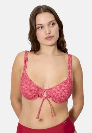 Joven mujer con un top de bikini rosa con estampado y detalle de lazo, mirando ligeramente hacia la derecha, sobre un fondo claro y liso.