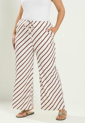 Frau trägt weiße Hose mit diagonalen braunen Streifen, ein weißes ärmelloses Oberteil und beige Sandalen mit dekorativen Riemen.