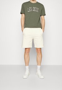 Oliwkowa bawełniana koszulka z nadrukiem "LES DEUX" w białym kolorze, zestawiona z kremowymi szortami i białymi trampkami.