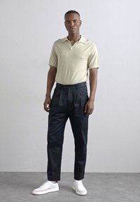 Lys beige kortermet strikket polo skjorte, svarte plisserte bukser og hvite joggesko. Glatte teksturer og skreddersydde designelementer.