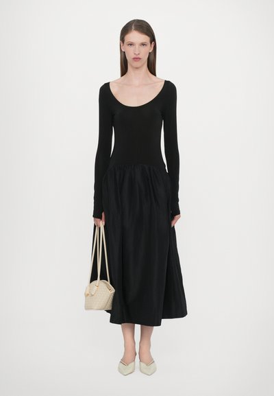 Vestido negro de manga larga con un corsé ajustado y falda fluida. Combinado con un bolso crema y zapatos de punta, de color claro.