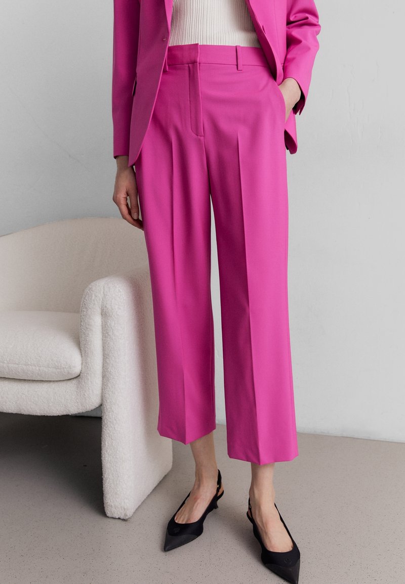 Femme portant un pantalon court taille haute rose vif, un blazer assorti, un haut blanc et des escarpins noirs à lanière arrière pointue, debout à côté d'un fauteuil blanc.