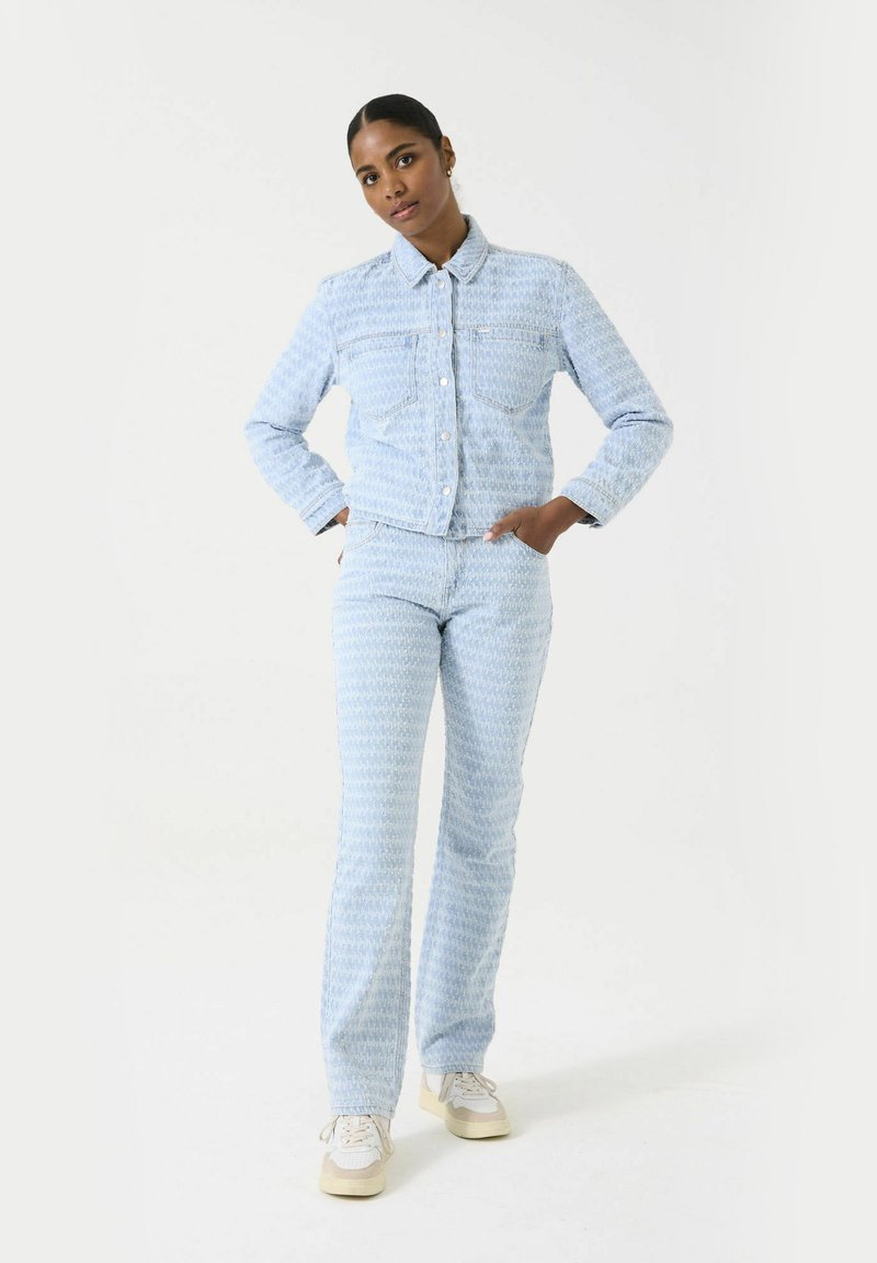 Lichtblauwe denimjack en bijpassende broek met een gestructureerd patroon. De outfit heeft een klassieke kraag en voorzakken.