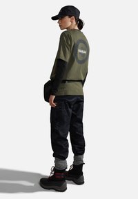 Maglietta verde oliva con un logo circolare sulla schiena, pantaloni cargo neri, calzini grigi e stivali neri con accenti rossi, indossando un berretto nero.