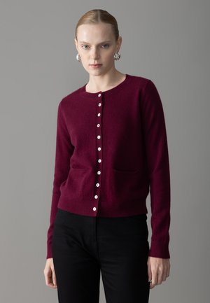 Burgundy cardigan valmistettuna pehmeästä materiaalista, jossa on pyöreä kaula-aukko, pitkät hihat ja valkoinen nappisuljenta sekä kaksi etutaskua.