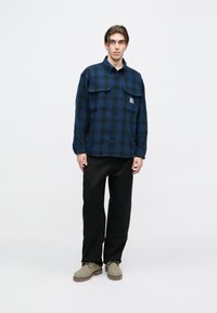 Πουκάμισο με κουμπιά σε ναυτικό μπλε και μαύρο καρό σχέδιο, διαθέτει δύο τσέπες στο στήθος και μια μικρή ετικέτα λογότυπου. Συνδυάζεται με μαύρο παντελόνι και μπεζ παπούτσια.