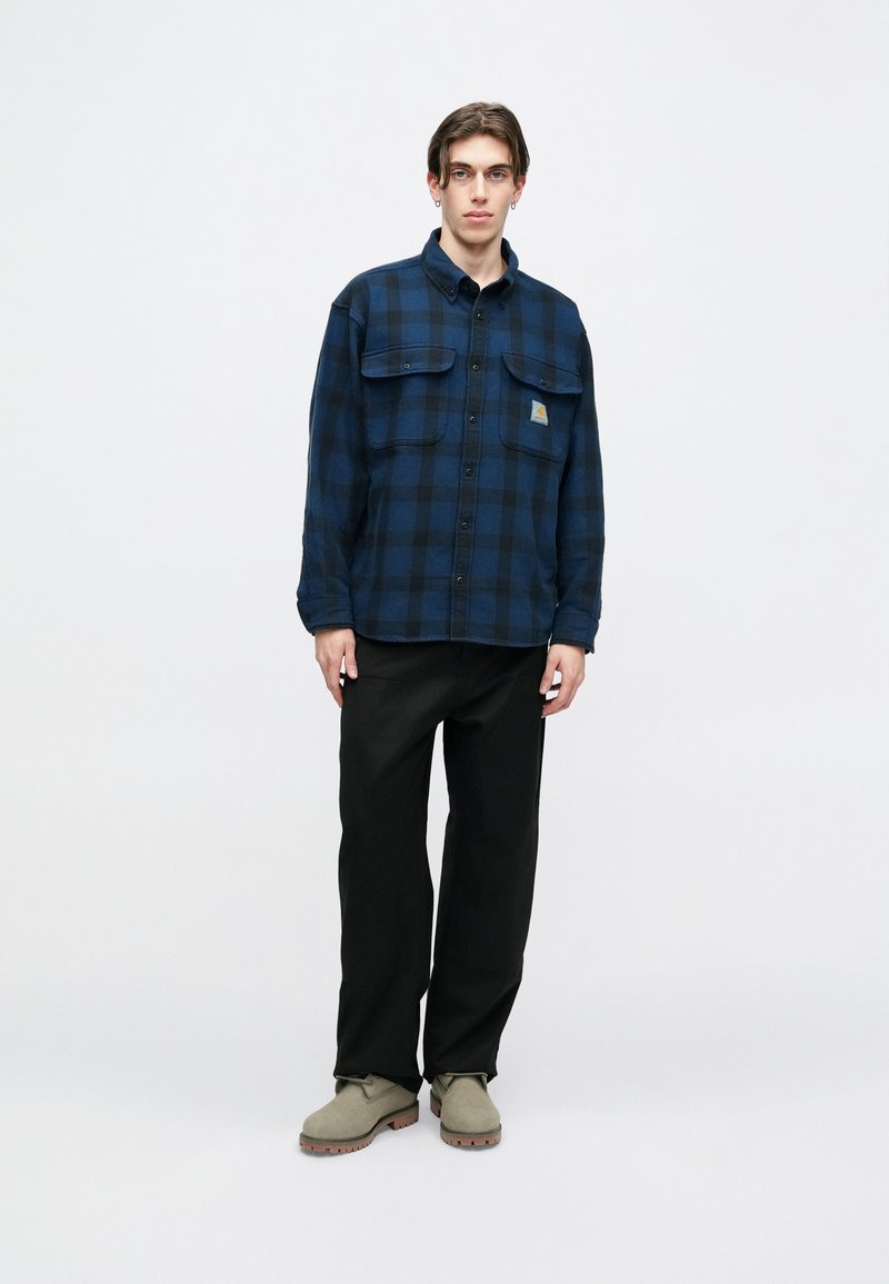 Overhemd met knopen in marineblauw en zwart geruit patroon, met twee borstzakken en een klein logo-opdruk; gecombineerd met zwarte broek en beige schoenen.