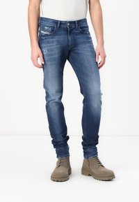 Μαύρες τζιν παντελόνες από κανονικό ύφασμα με στενή εφαρμογή, που διαθέτουν ελαφρές φθορές, πέντε τσέπες και κλείσιμο με κουμπί. Φοριούνται με καφέ δερμάτινα μποτάκια με κορδόνια.