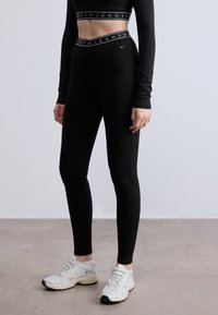 Μαύρο τοπ με μακριά μανίκια και λάστιχο με λογότυπο, συνδυασμένο με μαύρο κολάν ψηλόμεσο. Το μοντέλο φοράει ανοιχτόχρωμα αθλητικά παπούτσια με υφασμάτινες λεπτομέρειες.