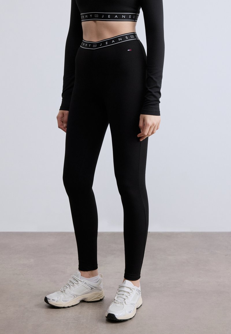 Μαύρο τοπ με μακριά μανίκια και λάστιχο με λογότυπο, συνδυασμένο με μαύρο κολάν ψηλόμεσο. Το μοντέλο φοράει ανοιχτόχρωμα αθλητικά παπούτσια με υφασμάτινες λεπτομέρειες.