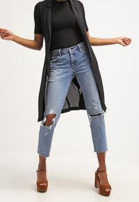 Person, der hellblaue, zerrissene Jeans, eine schwarze, ärmellose lange Weste, ein schwarzes, kurzärmliges Oberteil und braune Plateauschuhe trägt, steht mit leicht erhobenen Armen.