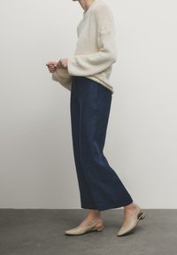 Beżowy sweter dzianinowy z szerokimi rękawami, w połączeniu z ciemnoniebieskimi spodniami o szerokim kroju i jasnobeżowymi butami na obcasie z odkrytymi palcami. Teksturowane materiały, luźny krój.