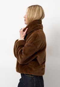 Bruine nepbontjas met een hoge kraag, voorzien van een zachte textuur en een cropped ontwerp. Gemaakt in combinatie met donkere denimspijkerbroeken.