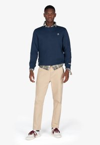 Un joven está de pie vistiendo un sudadera navy sobre una camisa de cuadros beige, pantalones beige, zapatillas blancas con cordones color burdeos y una pulsera en la muñeca.