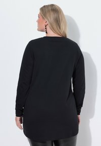 Schwarzes Langarmshirt mit geradem Schnitt und weichem Stoff, ausgestattet mit einem runden Halsausschnitt und verlängertem hinteren Saum. Sichtbare Seitennähte und Bündchen.