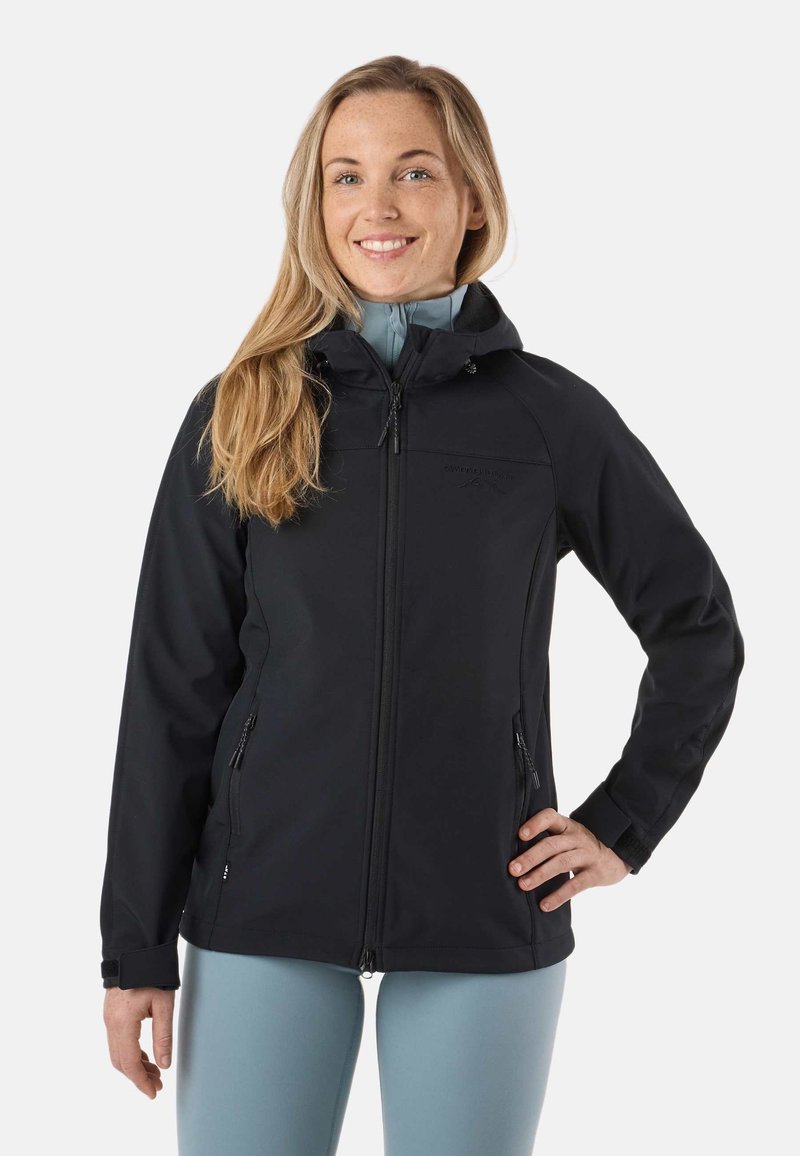 Femme souriante aux longs cheveux blonds portant une veste noire à capuche avec fermeture éclair et des leggings bleu clair, debout, une main sur la hanche.