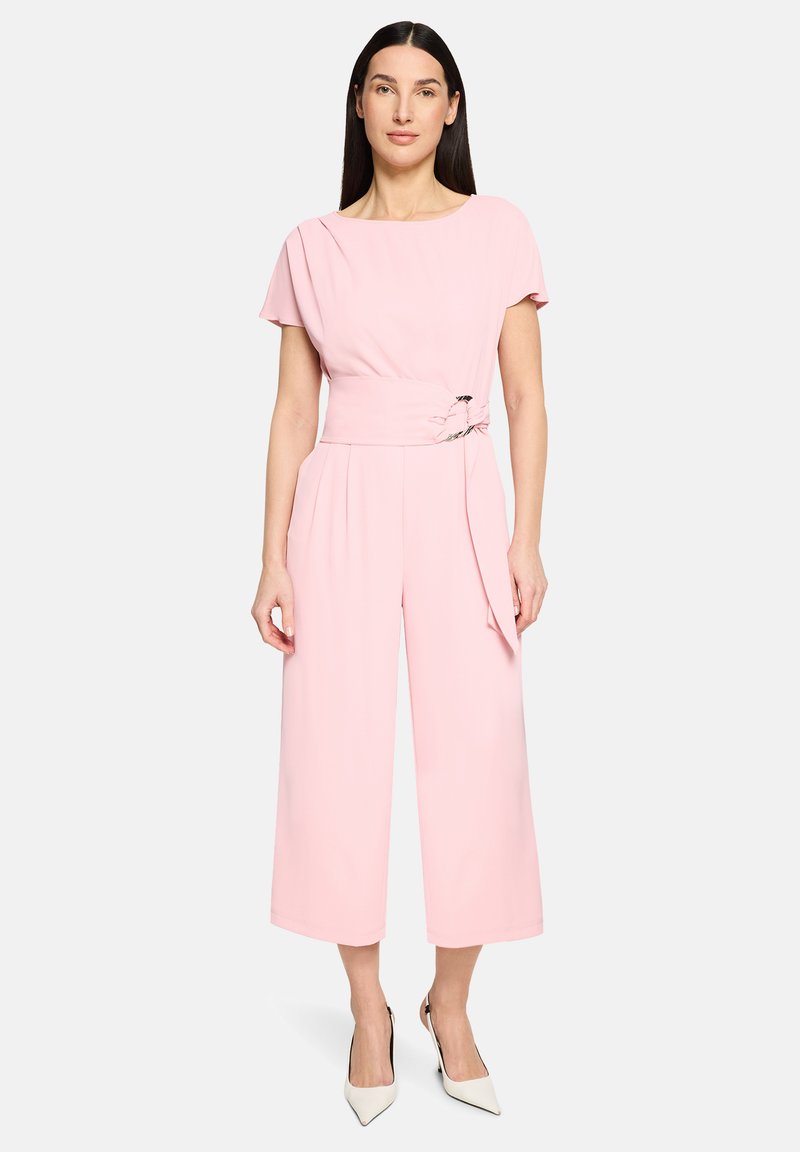 Femme debout portant une combinaison rose clair à manches courtes avec une ceinture large et des talons pointus blancs, sur un fond blanc uni.