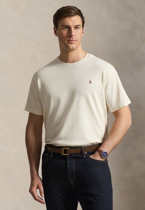 Man wearing off-white short-sleeve shirt tucked into dark jeans with brown belt, hand in pocket, and blue-faced wristwatch.