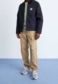 Marineblaue Canvas-Jacke mit Reißverschluss, khakifarbene Cargohose und graue Turnschuhe mit blauen Akzenten. Das Model trägt ein grünes Hemd unter der Jacke.