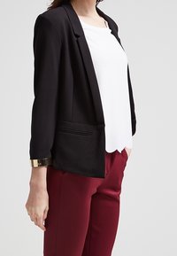 Blazer noir avec un design ajusté, en tissu léger, et des poignets accentués de doré, associé à un haut blanc et un pantalon bordeaux.