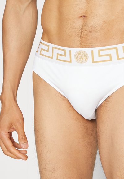 Weiße Bikini-Brief mit goldenem griechischem Schlüssel-Muster am Bund und Logo-Akzent. Weicher, glatter Stoff mit enger Passform und minimaler Bedeckung.