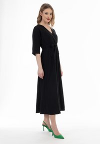 Sort omslagskjole med trekvartærmer og bælte i taljen, parret med grønne slingback hæle. Glat stof og ankel-længde design.
