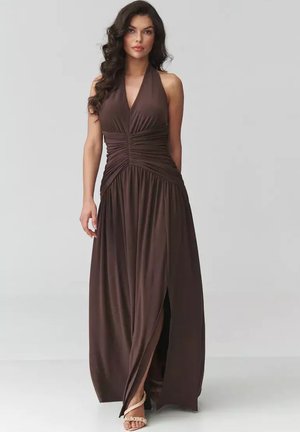 Kobieta z długimi ciemnymi włosami w brązowej, bezrękawnikowej sukience maxi z dekoltem typu halter, marszczoną talią i rozcięciem z przodu, stojąca na szarym tle.