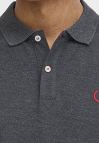 Muž v tmavě šedé texturované polo košili s bílými knoflíky a červeným vyšitým logem na hrudi.