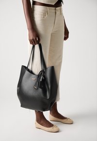 Persoon in lichtbeige jeans en crèmekleurige ballerina's die een grote zwarte leren Guess-tas aan de handvatten vasthoudt.