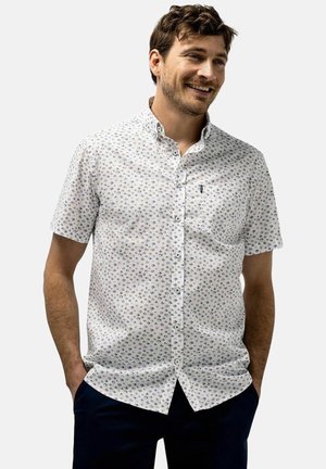 Man smiling with hands in pockets wearing a white short-sleeve button-up shirt with small dark patterns and dark pants.