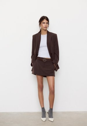 Mladá žena stojaca opretá o bielu stenu, oblečená v hnedom pruhovanom blejzri a sukni, bielom topu, sivých ponožkách a bielych lodičkách.